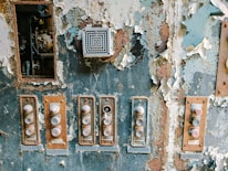 A weathered and rusty control panel with peeling paint and multiple buttons that appear worn. The buttons are arranged in a vertical fashion with labels like 'START'. A small vent or speaker is visible above the buttons. The surrounding paint is flaking off, revealing rust underneath.