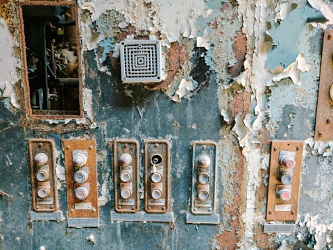 A weathered and rusty control panel with peeling paint and multiple buttons that appear worn. The buttons are arranged in a vertical fashion with labels like 'START'. A small vent or speaker is visible above the buttons. The surrounding paint is flaking off, revealing rust underneath.