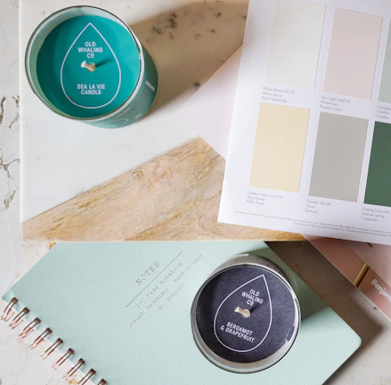 A serene flat lay of a cream journal, gold pen, and a softly glowing candle on a white surface.