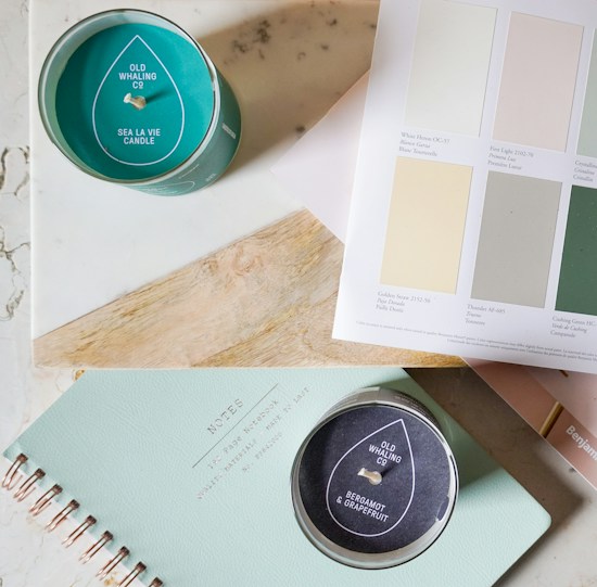 A flat lay of a candle, a notebook, and a color palette. The candle has a teal color with a label that reads 'Old Whaling Co, Sea La Vie Candle.' Next to it, a black-labeled candle with 'Bergamot & Grapefruit' written on it. A spiral-bound notebook with 'NOTES' embossed on the cover is also visible. Above the notebook, there is a color palette with various pastel shades and their names.