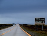 A long, empty road stretches into the distance under a cloudy, gray sky. On the right side, a green road sign displays the distances to Puerto Natales and Punta Arenas, along with the words 'Ruta del Fin del Mundo' and 'Circuito Aonikenk.' Two cars are visible further down the road, adding a sense of scale and distance.