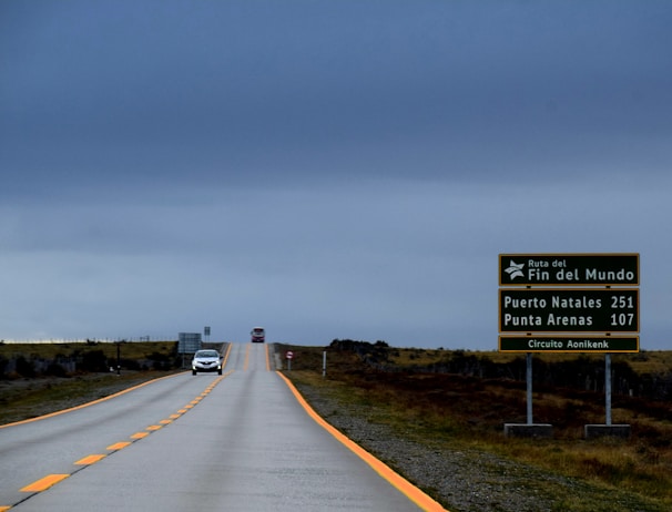 A long, empty road stretches into the distance under a cloudy, gray sky. On the right side, a green road sign displays the distances to Puerto Natales and Punta Arenas, along with the words 'Ruta del Fin del Mundo' and 'Circuito Aonikenk.' Two cars are visible further down the road, adding a sense of scale and distance.