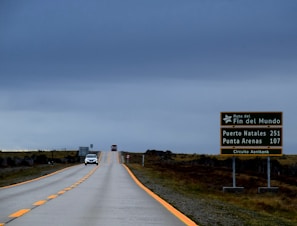 A long, empty road stretches into the distance under a cloudy, gray sky. On the right side, a green road sign displays the distances to Puerto Natales and Punta Arenas, along with the words 'Ruta del Fin del Mundo' and 'Circuito Aonikenk.' Two cars are visible further down the road, adding a sense of scale and distance.