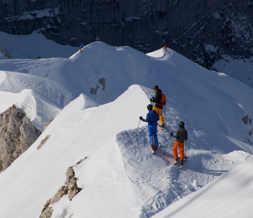 A group of skiers is navigating a snow-covered mountain ridge. The snow is pristine, with ski tracks visible on the surface. The skiers are wearing colorful outfits, adding contrast to the white snow. The background features rugged, rocky cliffs.