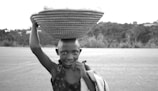 A young person carrying a large woven basket on their head stands in the foreground, smiling and looking directly at the camera. The background features a blurry landscape with trees and open space, suggesting a rural setting.
