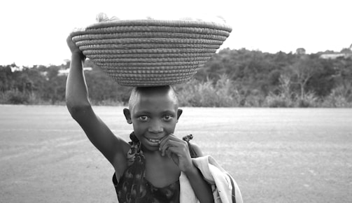 A young person carrying a large woven basket on their head stands in the foreground, smiling and looking directly at the camera. The background features a blurry landscape with trees and open space, suggesting a rural setting.
