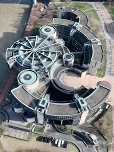 An aerial view of a large, modern architectural complex featuring circular and semi-circular buildings with gray roofs. The structure is distinctive, resembling a rotary shape with multiple arms extending from a central point. The surrounding area includes pathways, some greenery, and a riverbank nearby, while several vehicles are parked along a curved road.
