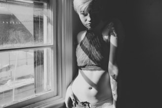 A black and white photograph of a person with short hair leaning against a sunlit window. The light casts shadows creating a dramatic effect on their body, revealing tattoos on their arm. They are wearing a patterned top and jeans, with a contemplative expression.