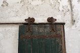 A rustic wooden door with a faded green color and metal hinges is mounted on a textured, peeling white wall. The door has a set of industrial-looking metal fixtures that suggest it slides to open. Above the door, there is a set of numbers painted directly on the wall.