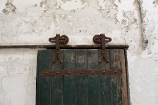 A rustic wooden door with a faded green color and metal hinges is mounted on a textured, peeling white wall. The door has a set of industrial-looking metal fixtures that suggest it slides to open. Above the door, there is a set of numbers painted directly on the wall.
