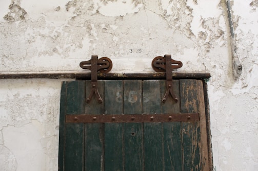 A rustic wooden door with a faded green color and metal hinges is mounted on a textured, peeling white wall. The door has a set of industrial-looking metal fixtures that suggest it slides to open. Above the door, there is a set of numbers painted directly on the wall.