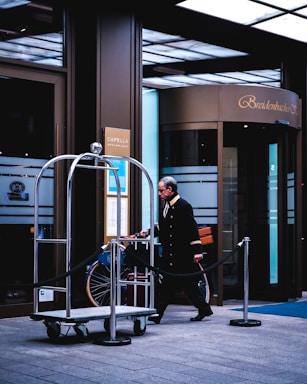 A hotel entrance scene features a uniformed doorman or bellhop walking by an empty luggage cart with a rope barrier alongside. The building has large glass doors with 'Breidenbacher Hof' displayed above the entrance. Signs for a bar and spa lounge are visible on the walls.