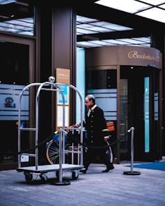 A hotel entrance scene features a uniformed doorman or bellhop walking by an empty luggage cart with a rope barrier alongside. The building has large glass doors with 'Breidenbacher Hof' displayed above the entrance. Signs for a bar and spa lounge are visible on the walls.