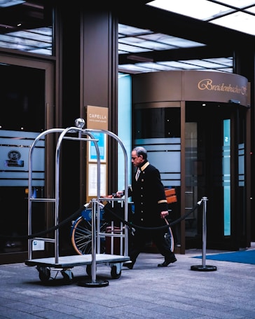 A hotel entrance scene features a uniformed doorman or bellhop walking by an empty luggage cart with a rope barrier alongside. The building has large glass doors with 'Breidenbacher Hof' displayed above the entrance. Signs for a bar and spa lounge are visible on the walls.
