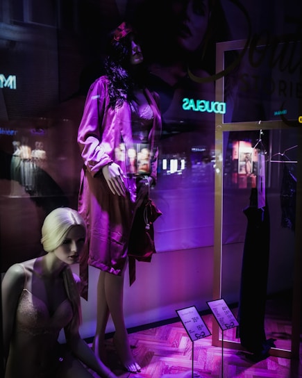 A display window featuring mannequins in a clothing store. One mannequin with dark hair is dressed in a pink satin robe, while another blonde mannequin is seated wearing pink lingerie. The window has a purple and dark ambiance with some reflections of the city lights visible. A black dress is hung on a gold frame, and there are price tags indicating discounts.