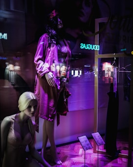 A display window featuring mannequins in a clothing store. One mannequin with dark hair is dressed in a pink satin robe, while another blonde mannequin is seated wearing pink lingerie. The window has a purple and dark ambiance with some reflections of the city lights visible. A black dress is hung on a gold frame, and there are price tags indicating discounts.