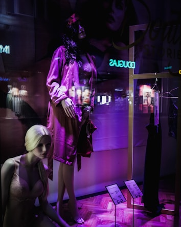 A display window featuring mannequins in a clothing store. One mannequin with dark hair is dressed in a pink satin robe, while another blonde mannequin is seated wearing pink lingerie. The window has a purple and dark ambiance with some reflections of the city lights visible. A black dress is hung on a gold frame, and there are price tags indicating discounts.