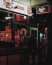 A dimly lit street scene featuring a noodle shop with signage in both English and Asian characters. A person wearing a dark coat is closely examining the menu displayed outside the shop, while another person in a padded jacket walks by. The shop window is covered with colorful posters promoting various dishes.