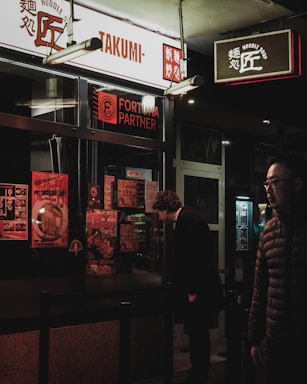 A dimly lit street scene featuring a noodle shop with signage in both English and Asian characters. A person wearing a dark coat is closely examining the menu displayed outside the shop, while another person in a padded jacket walks by. The shop window is covered with colorful posters promoting various dishes.