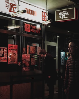 A dimly lit street scene featuring a noodle shop with signage in both English and Asian characters. A person wearing a dark coat is closely examining the menu displayed outside the shop, while another person in a padded jacket walks by. The shop window is covered with colorful posters promoting various dishes.