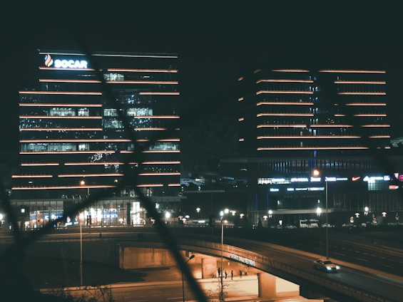 Several modern office buildings are illuminated at night, featuring bright horizontal lines of neon lights. A highway runs in the foreground, with a few vehicles visible. The buildings exhibit a sleek architectural design, with one displaying the SOCAR logo.