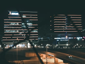 Several modern office buildings are illuminated at night, featuring bright horizontal lines of neon lights. A highway runs in the foreground, with a few vehicles visible. The buildings exhibit a sleek architectural design, with one displaying the SOCAR logo.