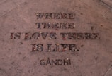 The image features an engraved quote on a stone surface, filled with small colorful stones and leaves. The quote says 'Where there is love there is life.' with 'Gandhi' attributed at the bottom.