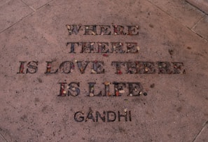 The image features an engraved quote on a stone surface, filled with small colorful stones and leaves. The quote says 'Where there is love there is life.' with 'Gandhi' attributed at the bottom.