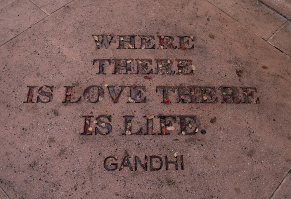 The image features an engraved quote on a stone surface, filled with small colorful stones and leaves. The quote says 'Where there is love there is life.' with 'Gandhi' attributed at the bottom.