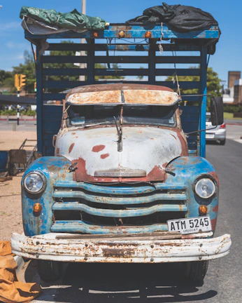 A vintage, rusted Chevrolet truck with a weathered blue and white exterior stands parked. The vehicle shows signs of age with significant rust and faded paint. The front license plate reads 'ATM 5245' from Uruguay. Various items, including a tarp, are piled on top. The scene is set in an urban area with some greenery and road signs visible in the background.