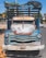 A vintage, rusted Chevrolet truck with a weathered blue and white exterior stands parked. The vehicle shows signs of age with significant rust and faded paint. The front license plate reads 'ATM 5245' from Uruguay. Various items, including a tarp, are piled on top. The scene is set in an urban area with some greenery and road signs visible in the background.