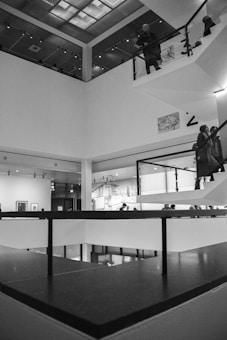An interior view of a modern, multi-level building. The scene shows several people standing on different floors, with a focus on sleek architectural elements like glass railings and white walls. Artworks are displayed on the walls, and soft, artificial lighting illuminates the space.