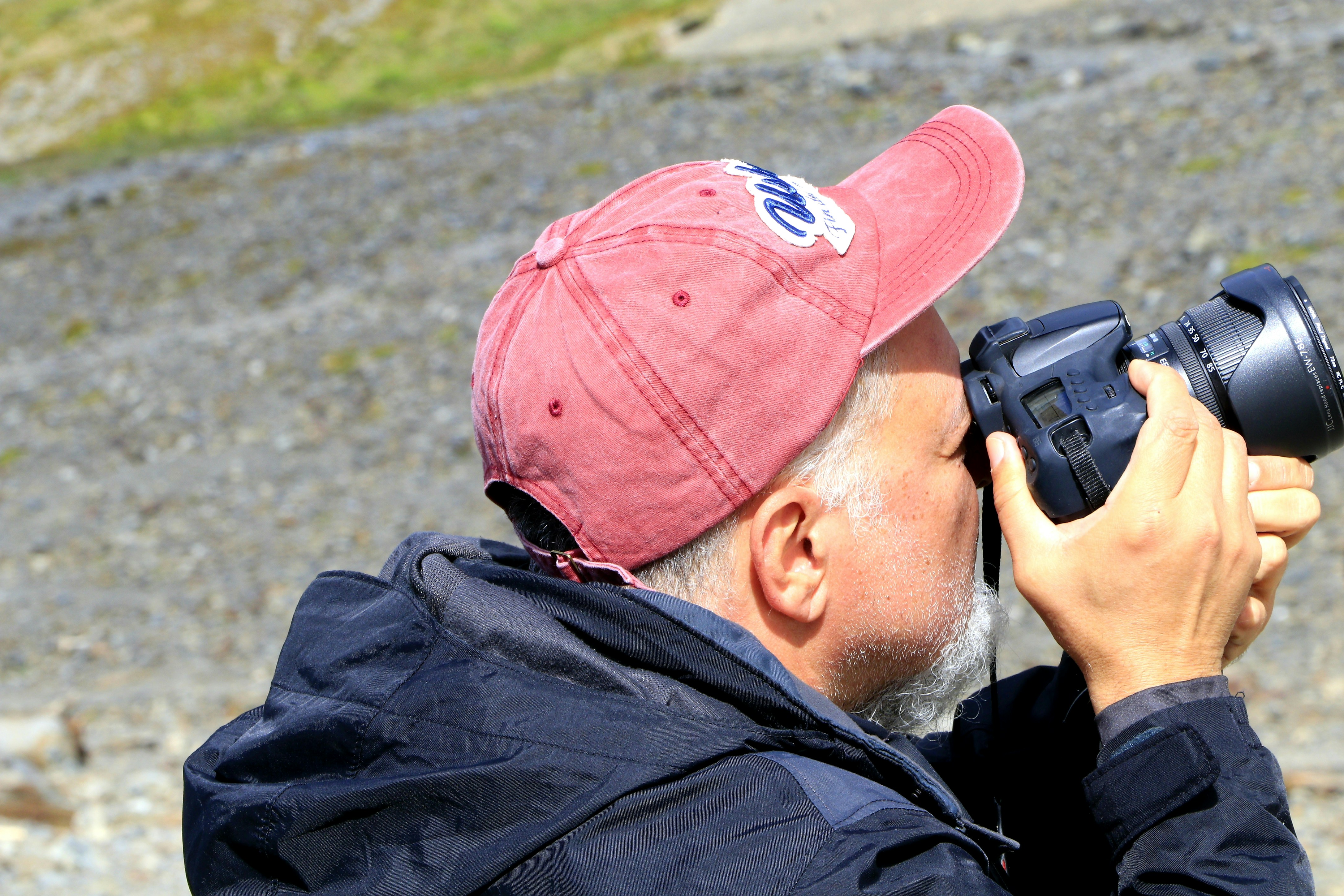 Photograph of a man in a red cap holding a DSLR up to his eye in a gravel outdoor setting.
