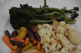 A white plate holds a selection of roasted vegetables, including broccolini, cauliflower, and various colors of carrots.