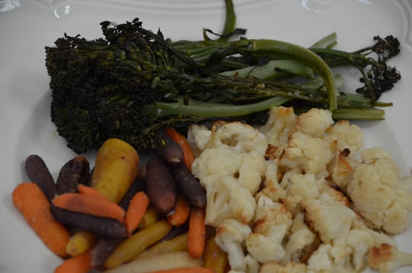 A white plate holds a selection of roasted vegetables, including broccolini, cauliflower, and various colors of carrots.