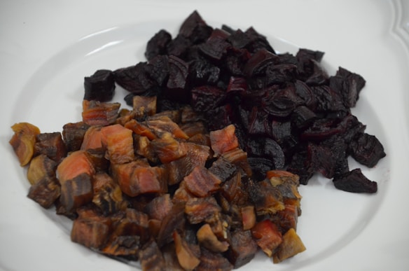 A white plate holding two distinct piles of diced food, one with dark brownish-red pieces and another with deep, dark purple pieces. The texture of the pieces looks cooked and somewhat glossy, indicative of roasted or baked preparation.