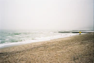 A foggy beach scene with overcast skies, gentle waves approaching the sandy shore, and a solitary person in a bright yellow jacket walking along the beach. Several seagulls are flying above the water, and there are concrete wave breakers in the distance.