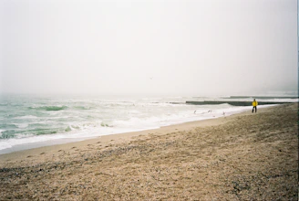 A foggy beach scene with overcast skies, gentle waves approaching the sandy shore, and a solitary person in a bright yellow jacket walking along the beach. Several seagulls are flying above the water, and there are concrete wave breakers in the distance.