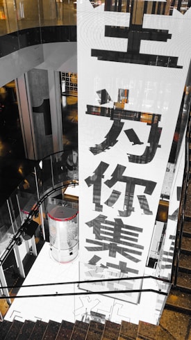 A large vertical banner with bold Chinese characters is displayed, dominating the interior space of a modern building. The architecture features dark reflective surfaces and a combination of industrial and sleek design elements.