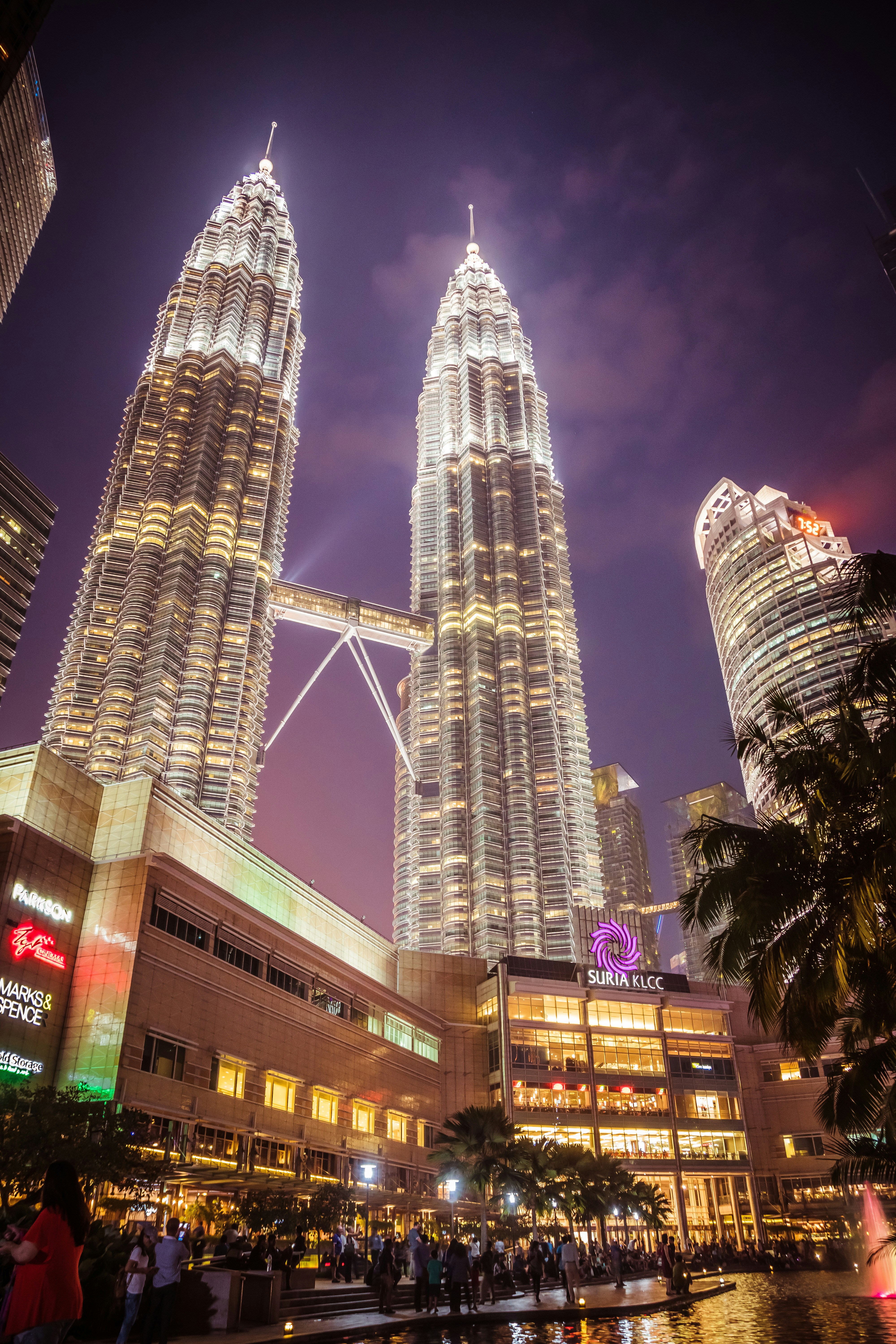 Kuala Lumpur Petronas Towers at dusk, gleaming steel facades, purple sunset sky, lush tropical greenery below