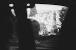 A black and white photograph depicts a cityscape with a long street lined by buildings and trees extending into the distance. The perspective appears to be from behind a dark object in the foreground, creating a frame for the bustling city view ahead. Sparse vegetation and distant hills are visible under the clear sky.