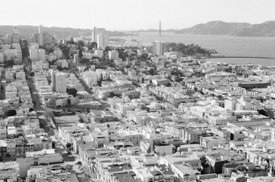 An aerial view of a densely packed urban area with numerous buildings and streets laid out in a grid pattern. In the background, a suspension bridge spans a body of water, with hills visible on the horizon. The image is in black and white, highlighting the architectural details and city layout.