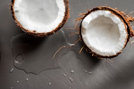 Two halves of a coconut with their white flesh exposed are placed on a grey textured surface. Water droplets and splashes are visible around the coconuts, suggesting freshness or recent cutting.