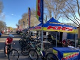 A row of bicycles is displayed under branded tents set up along a road. Several people are interacting, with one child riding a bicycle in the foreground. The trees surrounding the area have bare branches, and the scene is illuminated by daylight, suggesting a clear day.