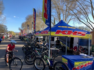 A row of bicycles is displayed under branded tents set up along a road. Several people are interacting, with one child riding a bicycle in the foreground. The trees surrounding the area have bare branches, and the scene is illuminated by daylight, suggesting a clear day.