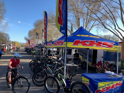 A row of bicycles is displayed under branded tents set up along a road. Several people are interacting, with one child riding a bicycle in the foreground. The trees surrounding the area have bare branches, and the scene is illuminated by daylight, suggesting a clear day.