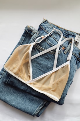 A sheer beige bralette with white trim is laid neatly on a pair of blue denim jeans. The jeans have a noticeable texture and stitching, and the bralette's delicate fabric adds a contrast of softness.