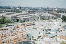 A busy amusement park scene featuring a roller coaster, various small buildings, and numerous people. In the background, there are several large industrial buildings and streetcars. The park area is lively and crowded, suggesting a popular attraction.