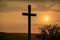 A wooden cross stands prominently in the foreground against a sunset sky. The sun is low, casting warm hues across the landscape, while clouds partially obscure the view above. In the lower right corner, silhouetted foliage adds a natural element to the scene.