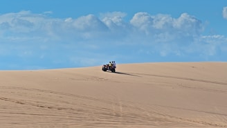 A person rides an ATV across expansive sand dunes under a vivid blue sky filled with scattered white clouds. The smooth sand has visible tire tracks, indicative of recent activity.